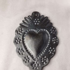 Könnte beinhalten: Schwarzes Metallerz in Herzform mit gewelltem Rand und floralen Designs. Das Herz ist erhaben und hat einen perlenbesetzten Rand.