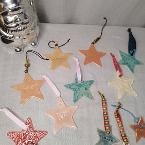 Può includere: Una collezione di ornamenti a forma di stella in ceramica in vari colori, tra cui pesca, bianco e verde acqua. Ogni stella ha un design diverso ed è infilata con un nastro o un cordoncino. Alcune stelle hanno parole come "Loved" e "Happy". Sullo sfondo una statuina di gnomo argentato.