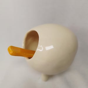 Peut inclure: Un pot à sel en céramique crème, de forme arrondie, avec trois petits pieds. Une cuillère jaune-orangé est insérée dans l'ouverture. Le pot à sel a une finition lisse et brillante.