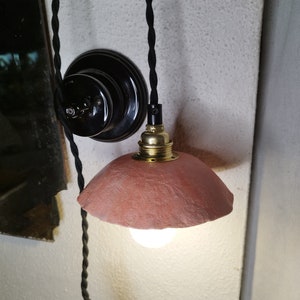 Peut inclure: Une petite lampe suspendue de couleur terre cuite avec un abat-jour texturé. La lampe est suspendue à un cordon torsadé noir et comporte un support d'ampoule de couleur laiton. Un interrupteur noir est visible sur le mur.