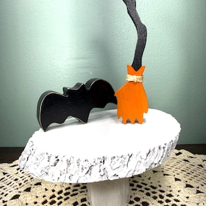 Puede incluir: Un murciélago de madera negra y una escoba en miniatura con mango negro y cerdas naranjas atadas con cuerda, exhibidos sobre un pedestal de madera pintado de blanco con borde de corteza natural y base cilíndrica.