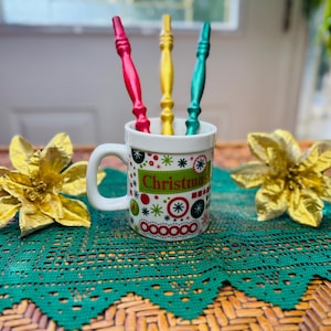 May include: A white mug with a Christmas design, holding three colourful stirrers. The mug is decorated with red, green, and white patterns and the word "Christmas". Two gold poinsettias and a green lace doily complete the festive scene.