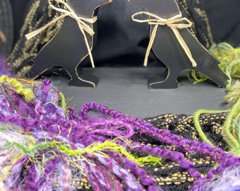 May include: Two black wooden crow figurines with a rustic finish. Each crow has a brown raffia bow tied around its neck. The crows are sitting on a black surface with purple and green yarn in the foreground.