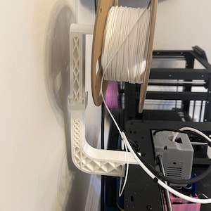 May include: White 3D printed filament holder mounted on a wall with a spool of white filament. The holder has a lattice design and a curved arm. The filament is attached to a 3D printer.