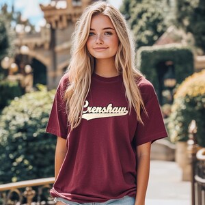 May include: A maroon t-shirt with the word "Crenshaw" in a white and gold script across the chest. The shirt has short sleeves and a crew neck.