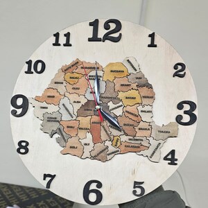 Może przedstawiać: Drewniany zegar z mapą Rumunii na tarczy. Tarcza zegara jest biała z czarnymi cyframi i wskazówkami. Mapa składa się z różnych kolorowych drewnianych elementów, z których każdy reprezentuje inny powiat w Rumunii. Powiaty są oznaczone nazwami w czarnym kolorze.
