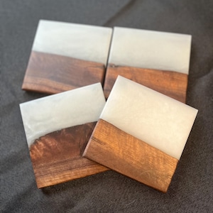 May include: Four square coasters with a two-tone design. The top half is a light, pearlescent color, and the bottom half is a rich, dark wood. The coasters are arranged in a slightly offset square.