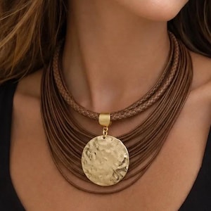 May include: A multi-strand brown leather cord necklace featuring a central gold-toned circular pendant with a hammered, textured surface. The necklace is secured with a gold-toned metal connector piece that holds the various leather strands together.