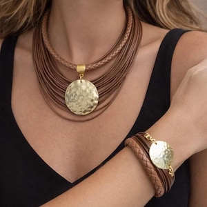 May include: A multi-strand necklace and matching bracelet set featuring brown leather cords, including braided and smooth strands, accented with a large, circular, gold-toned hammered metal pendant and a smaller matching circular charm.