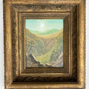 Puede incluir: Una pintura de paisaje antigua en un marco dorado y ornamentado. La obra de arte representa un valle con una cascada, montañas y un brillante estallido de sol en el cielo. El marco tiene un diseño detallado de vid.