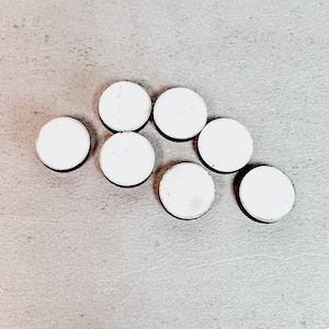 May include: Seven small, round, white objects with a silver-colored edge are arranged on a gray surface. The objects are evenly spaced and appear to be made of a smooth, reflective material. The overall composition is simple and clean.