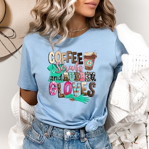 May include: A light blue t-shirt with the text "Coffee, Scrubs and Rubber Gloves" in colorful letters. The design includes a coffee cup with a medical cross, a stethoscope, and a pair of blue rubber gloves.