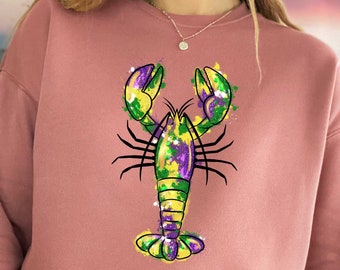 May include: A pink sweatshirt with a green, yellow, and purple lobster design. The lobster is painted in a Mardi Gras style.