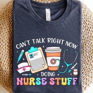 May include: Dark gray t-shirt with the text "Can't talk right now doing nurse stuff" in colorful letters. The design includes a stethoscope, a coffee cup, a thermometer, a syringe, a pill bottle, and a nurse's hat.