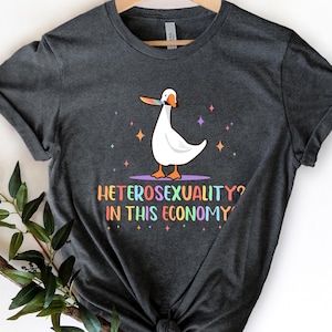 May include: A dark gray t-shirt with a white cartoon goose holding a knife in its beak. The text "Heterosexuality? In this economy?" is printed in rainbow colors below the goose.