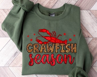 Könnte beinhalten: Grüner Pullover mit einem roten Krebs-Motiv und dem Text "Crawfish season" in einer Leopardenmuster-Schrift.