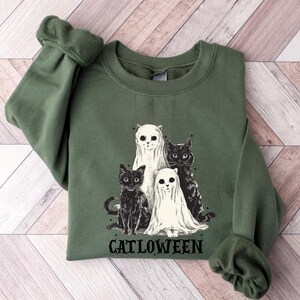 K&ouml;nnte beinhalten: Ein gr&uuml;ner Pullover mit einem schwarz-wei&szlig;en Motiv von drei Katzen, die als Geister verkleidet sind. Der Text "Catlloween" ist unter dem Motiv gedruckt.