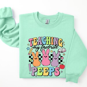 May include: A light green sweatshirt with the text "TEACHING my favorite PEEPS" in rainbow colors. The design includes three pastel-colored Peeps, a pencil, a book, a ruler, and an apple.