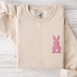 May include: A beige sweatshirt with a pink bunny rabbit applique on the front. The bunny has a white tail.