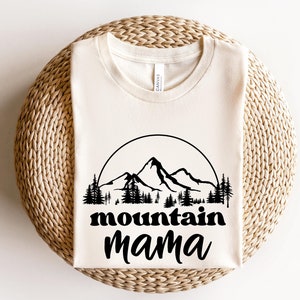 May include: Cream-colored t-shirt with a black mountain graphic inside a circle, with the words "mountain mama" in a bold font. The design includes mountain peaks and pine trees. The shirt is made of a soft material.