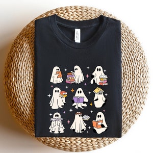 May include: Black t-shirt featuring a whimsical design of cartoon ghosts reading books. The ghosts are in various poses, holding and surrounded by colorful stacks of books. The design includes stars and a playful, book-themed aesthetic.