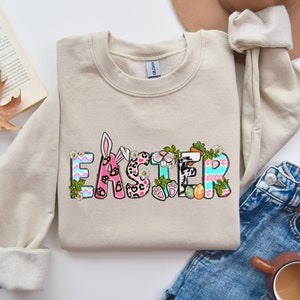 May include: A beige sweatshirt with the word "Easter" printed on the front in colorful letters. The letters are decorated with bunny ears, flowers, and polka dots.