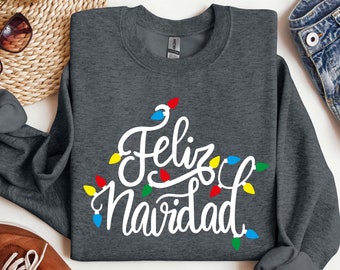 May include: A dark gray sweatshirt with the text "Feliz Navidad" in white lettering surrounded by colorful string lights. The sweatshirt is laid out on a white surface with other items around it.