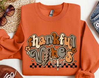 May include: Orange sweatshirt with a retro-style graphic that says "thankful vibes" in brown, yellow, and white lettering. The graphic also includes a checkered pattern and a stylized bone.