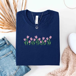 K&ouml;nnte beinhalten: Ein marineblaues T-Shirt mit rosa Tulpenstickerei. Eine silberne Halskette mit einem Sonnenanh&auml;nger liegt auf dem Shirt. Das Shirt wird mit blauen Jeans, wei&szlig;en Turnschuhen und einem beigen Schal kombiniert.