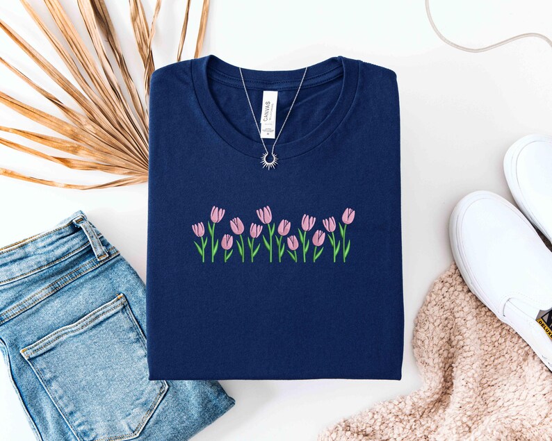K&ouml;nnte beinhalten: Ein marineblaues T-Shirt mit rosa Tulpenstickerei. Eine silberne Halskette mit einem Sonnenanh&auml;nger liegt auf dem Shirt. Das Shirt wird mit blauen Jeans, wei&szlig;en Turnschuhen und einem beigen Schal kombiniert.