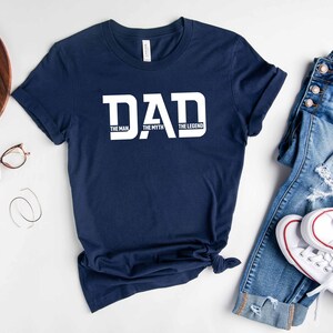 May include: A navy blue t-shirt with white text that reads "DAD THE MAN THE MYTH THE LEGEND". The shirt is laid flat on a white surface with a pair of jeans and white sneakers.