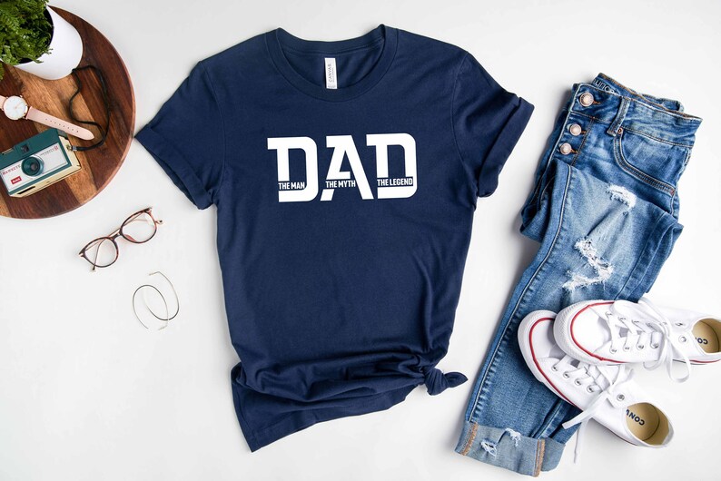 May include: A navy blue t-shirt with white text that reads "DAD THE MAN THE MYTH THE LEGEND". The shirt is laid flat on a white surface with a pair of jeans and white sneakers.