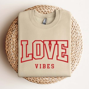 May include: A beige sweatshirt with the text "LOVE VIBES" in red lettering. The word "LOVE" is in a large, bold font with a red outline.