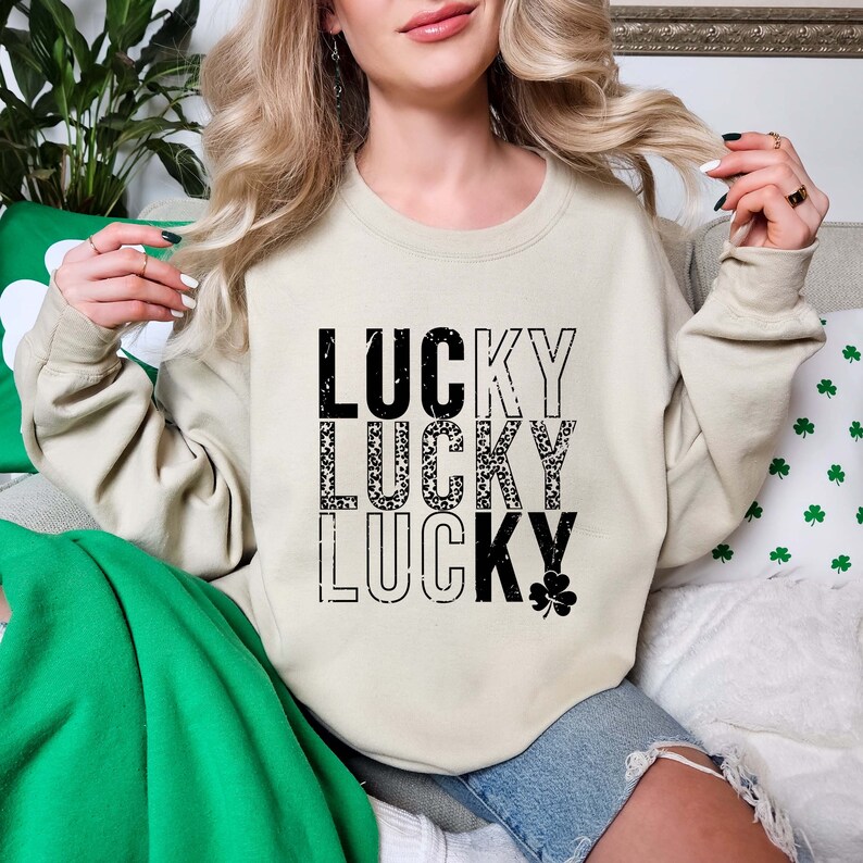 May include: A beige sweatshirt with the word "LUCKY" repeated three times in black text. The second "LUCKY" is printed with a black and white clover pattern. A small black clover is printed below the third "LUCKY".