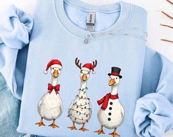 Może przedstawiać: Jasnoniebieska bluza z trzema gęsiami w świątecznych strojach. Jedna gęś ma czapkę Mikołaja i muszkę, druga ma rogi i lampki choinkowe, a trzecia jest przebrana za bałwana z cylindrem i szalikiem. Srebrny naszyjnik jest na bluzie.