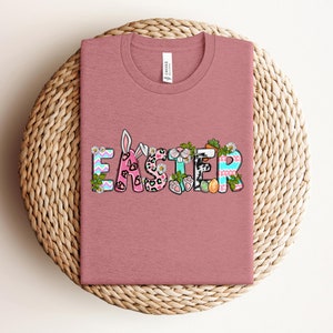 May include: A pink t-shirt with the word "Easter" spelled out in colorful letters with bunny ears, paw prints, and other Easter-themed designs.