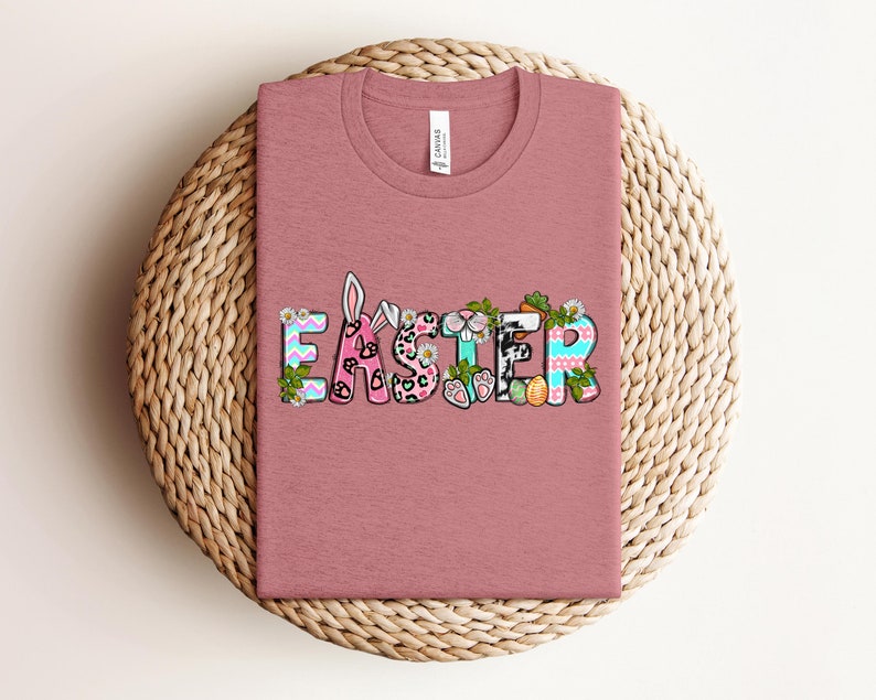 May include: A pink t-shirt with the word "Easter" spelled out in colorful letters with bunny ears, paw prints, and other Easter-themed designs.