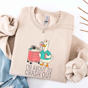 May include: Beige sweatshirt with a cartoon goose dressed as a nurse, holding a red can and standing next to a medical cart. The text "I'M ABOUT TO CRASH OUT" is printed below the goose. A silver necklace is draped across the sweatshirt.