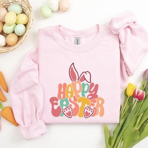 May include: Pink sweatshirt with a colorful "Happy Easter" graphic featuring a bunny rabbit. The graphic is in a retro style with a layered look.