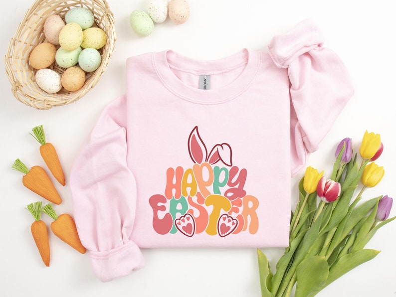 May include: Pink sweatshirt with a colorful "Happy Easter" graphic featuring a bunny rabbit. The graphic is in a retro style with a layered look.
