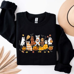 Może przedstawiać: Czarna bluza z nadrukiem graficznym kot&oacute;w przebranych na Halloween. Koty noszą kostiumy i trzymają dynie. Grafika jest w stylu kresk&oacute;wkowym.