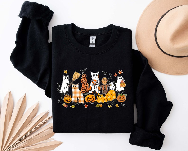 Może przedstawiać: Czarna bluza z nadrukiem graficznym kot&oacute;w przebranych na Halloween. Koty noszą kostiumy i trzymają dynie. Grafika jest w stylu kresk&oacute;wkowym.