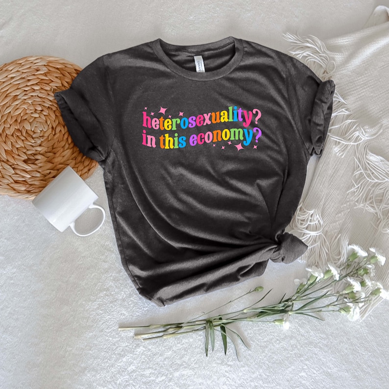 May include: A dark gray t-shirt with the text "heterosexuality? in this economy?" printed in rainbow colors.