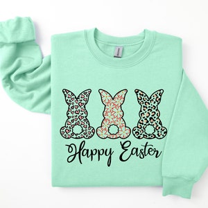 Puede incluir: Una sudadera de color verde claro con un gr&aacute;fico de tres conejos con diferentes patrones. Los conejos llevan patrones rosa, negro y naranja. El texto "Happy Easter" est&aacute; impreso debajo de los conejos.