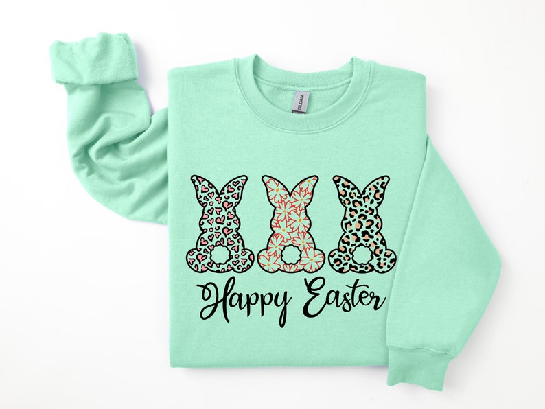 Puede incluir: Una sudadera de color verde claro con un gr&aacute;fico de tres conejos con diferentes patrones. Los conejos llevan patrones rosa, negro y naranja. El texto "Happy Easter" est&aacute; impreso debajo de los conejos.