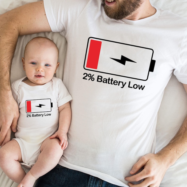 May include: A white t-shirt with a black and red battery icon and the text "2% Battery Low". A baby is wearing a smaller version of the same shirt.