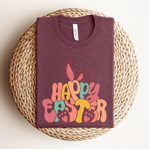 May include: A maroon t-shirt with a retro-style graphic that says "Happy Easter" in colorful letters. The graphic features a bunny rabbit with floppy ears.