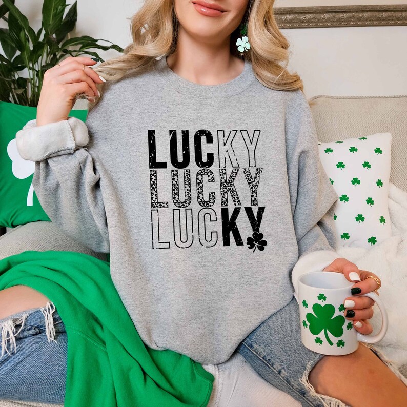 May include: A gray sweatshirt with the word "LUCKY" printed three times in black, with a shamrock graphic below the last word. The sweatshirt is being worn by a person sitting on a couch.