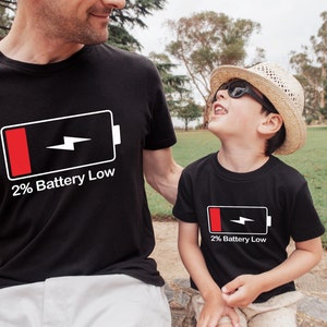 May include: Two people wearing black t-shirts with a white battery icon and the text "2% Battery Low". The person on the left is an adult and the person on the right is a child. The child is wearing a straw hat and sunglasses.