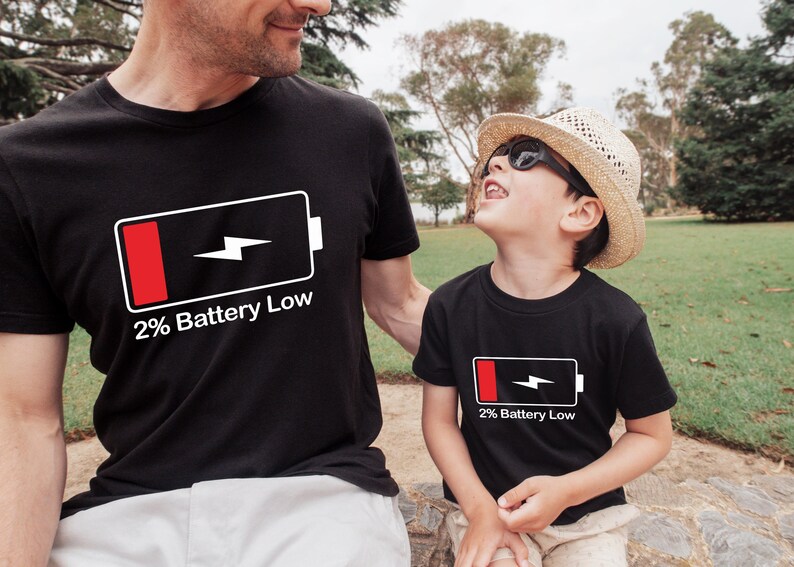 May include: Two people wearing black t-shirts with a white battery icon and the text "2% Battery Low". The person on the left is an adult and the person on the right is a child. The child is wearing a straw hat and sunglasses.